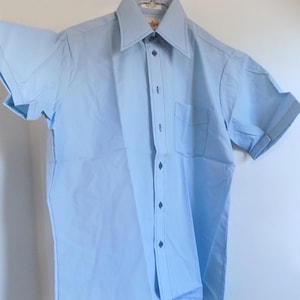 Camisa azul de manga curta, modelo deadstock, da década de 1970, com costuras decorativas, gola em papelão ainda presa à camisa.