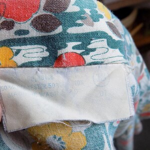 Puede incluir: Primer plano de una tela con un estampado floral colorido. La tela es de color verde azulado, amarillo, rojo y marr&oacute;n. Una etiqueta blanca est&aacute; cosida a la tela con un texto demasiado borroso para leer.