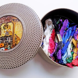 May include: A round, brown and white wicker-style box with a lid. The lid has a circular image of a girl drawing water from a well. The box is filled with colorful embroidery floss.