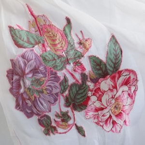 May include: A white fabric garment featuring a large floral applique with purple, pink, and green petals and leaves. The floral design is outlined with a bright pink stitched border, creating a textured, layered appearance on the white base.