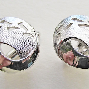 May include: A pair of silver cufflinks with a textured, circular design. Each cufflink features a cut-out pattern of leaves or branches.