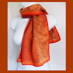 May include: A rectangular scarf featuring a vibrant orange and deep red colour palette with a repeating diagonal line pattern and small circular motifs. The scarf is finished with a solid orange border along the edges, creating a distinct frame.