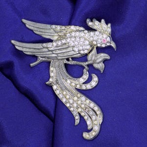 May include: Silver-toned bird brooch with clear and pink rhinestones. The bird is perched on a branch with a leaf, and has detailed wings and tail feathers. The brooch is set against a blue background.