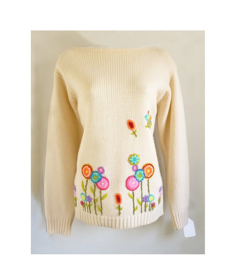 May include: A cream-colored knitted sweater with colorful embroidered flowers. The flowers are in shades of pink, blue, green, orange, and yellow.