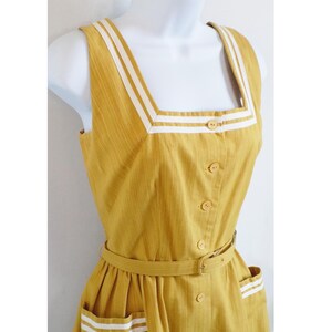 Pode incluir: Um vestido amarelo sem mangas com acabamento branco ao redor do decote e bolsos. O vestido tem um cinto e fecho de botões.