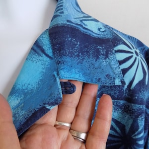 May include: Close-up of a blue and white patterned shirt collar with a button closure. The fabric has a textured, slightly distressed look.