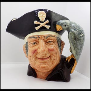 Può includere: Una tazza in ceramica a tema pirata. La tazza presenta un volto dettagliato con un cappello nero ornato da un teschio e ossa incrociate. Un pappagallo grigio è appollaiato sulla spalla. La tazza è dipinta in tonalità di marrone, nero e grigio.