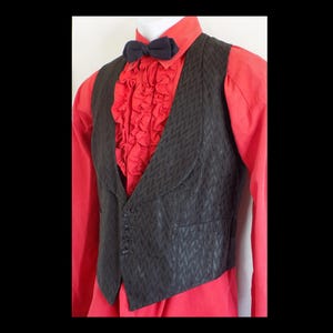Puede incluir: Un chaleco negro con un sutil patrón de espiga sobre una camisa roja con volantes. Una pajarita azul marino en el cuello. El chaleco tiene cierre de cuatro botones y un corte entallado, creando una estética vintage o teatral.