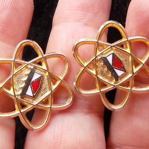 Puede incluir: Un par de gemelos de tono dorado con un diseño de átomo estilizado y un escudo rojo, blanco y negro en el centro.