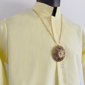 Könnte beinhalten: Ein gelbes Hemd mit Kragen, einer goldenen Kettenkette und einem großen, runden Anhänger mit türkisfarbenen und violetten Edelsteinen.
