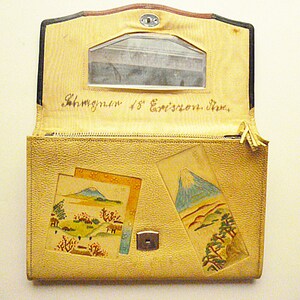 May include: A beige leather wallet with a zipper closure and a small mirror on the inside. The wallet has three decorative patches with hand-painted images of Mount Fuji. The wallet is inscribed with the text "Schwyzer 48' Exposition, Zurich."