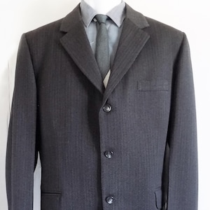 Könnte beinhalten: Ein dunkel anthrazitfarbener Blazer mit dezentem Fischgrätenmuster, drei Knöpfen auf der Vorderseite, einer Brusttasche und zwei unteren Pattentaschen. Der Blazer wird über einem hellgrauen Hemd mit Kragen und einer dunkelgrauen Krawatte getragen.