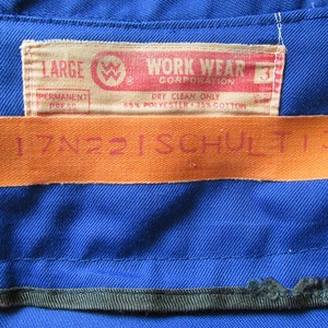 Puede incluir: Una camisa de trabajo azul con una etiqueta que dice "LARGE WORK WEAR CORPORATION" y "DRY CLEAN ONLY 65% POLYESTER - 35% COTTON". Una segunda etiqueta naranja dice "17N221SCHULT13".