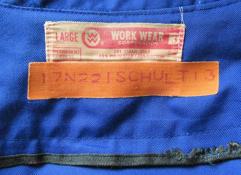 Puede incluir: Una camisa de trabajo azul con una etiqueta que dice "LARGE WORK WEAR CORPORATION" y "DRY CLEAN ONLY 65% POLYESTER - 35% COTTON". Una segunda etiqueta naranja dice "17N221SCHULT13".