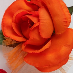 May include: A large, bright orange fabric rose with a tulle overlay. The rose has a layered, ruffled appearance and is accented with green fabric leaves.