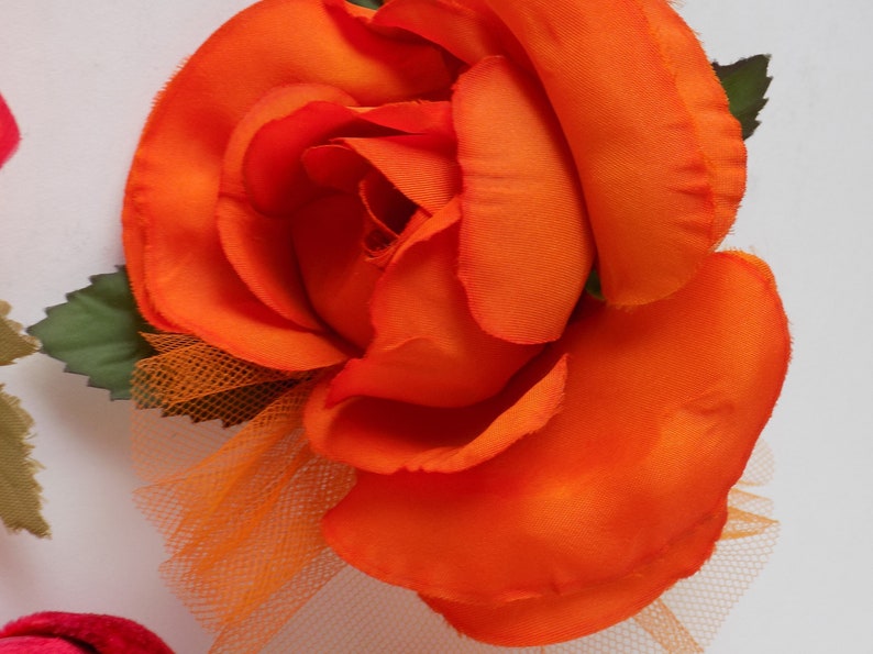 May include: A large, bright orange fabric rose with a tulle overlay. The rose has a layered, ruffled appearance and is accented with green fabric leaves.