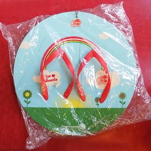 Puede incluir: Un letrero decorativo con forma de chancla con correas rojas y un arco iris, sobre un fondo de cielo azul con nubes y flores. El letrero tiene el texto "Force Good" y "New Beetle".