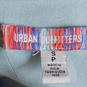 Puede incluir: Una etiqueta a rayas azul y blanca con el texto "URBAN OUTFITTERS" en rojo y blanco. La etiqueta tambi&eacute;n tiene una etiqueta blanca m&aacute;s peque&ntilde;a adjunta con la letra "S" y el texto "MADE IN INDIA, FABRIQU&Eacute; EN INDE".