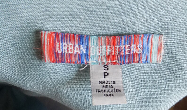 Puede incluir: Una etiqueta a rayas azul y blanca con el texto "URBAN OUTFITTERS" en rojo y blanco. La etiqueta tambi&eacute;n tiene una etiqueta blanca m&aacute;s peque&ntilde;a adjunta con la letra "S" y el texto "MADE IN INDIA, FABRIQU&Eacute; EN INDE".