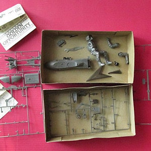 Puede incluir: Un kit de maquetas para un vehículo futurista y un piloto. El kit incluye piezas de plástico, instrucciones y una hoja de calcomanías. El vehículo es una nave espacial plateada elegante con una cabina y alas. El piloto es una figura plateada con un casco y un traje espacial. El kit está empaquetado en una caja de cartón con una imagen del modelo terminado en la parte delantera.