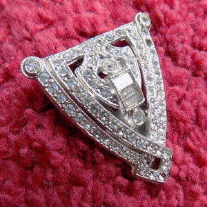 Puede incluir: Un broche de forma geométrica plateada con un diseño central de diamantes de imitación transparentes. El broche está rodeado por un borde de diamantes de imitación más pequeños.