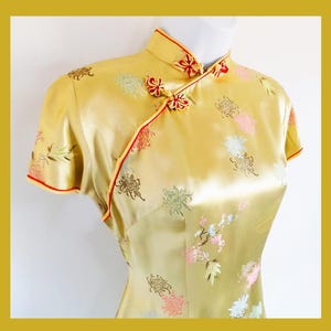 Puede incluir: Un cheongsam de seda dorado con cuello mandarín y ribetes rojos. El vestido está decorado con un estampado floral.