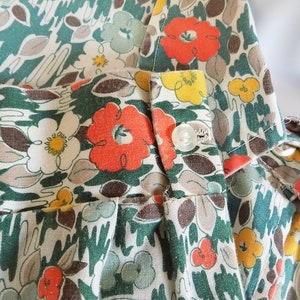 Puede incluir: Primer plano de una tela floral verde, blanca y naranja con un bot&oacute;n. La tela tiene un estilo vintage y es perfecta para proyectos de costura.