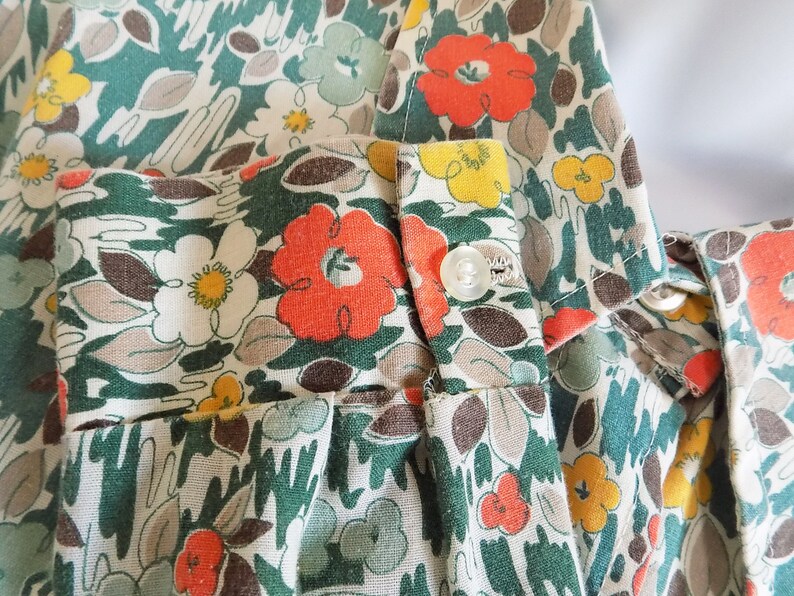 Puede incluir: Primer plano de una tela floral verde, blanca y naranja con un bot&oacute;n. La tela tiene un estilo vintage y es perfecta para proyectos de costura.