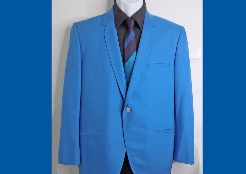 May include: A light blue blazer with a single button closure. The blazer has a notched lapel and a pocket on the left side. The blazer is worn over a dark gray shirt with a blue and white striped tie.