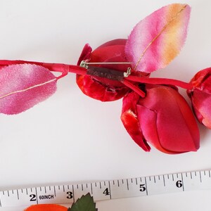 May include: A red velvet rose bud brooch with a pink and orange leaf. The brooch is attached to a brown stem with a silver pin.