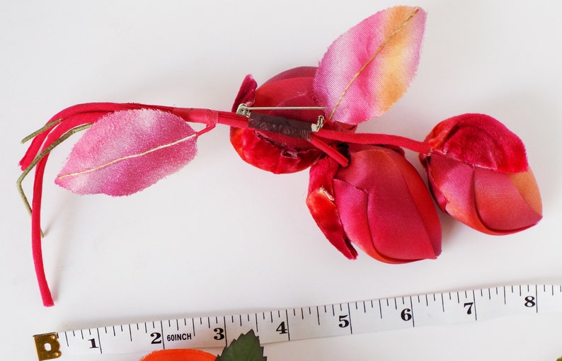 May include: A red velvet rose bud brooch with a pink and orange leaf. The brooch is attached to a brown stem with a silver pin.