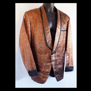 Könnte beinhalten: Ein kupferfarbener Blazer mit einem schwarzen Satin-Schal-Kragen und Manschetten. Der Blazer hat eine strukturierte, fast metallische Oberfläche.