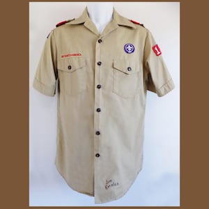 May include: Tan short-sleeved Boy Scouts of America shirt with a button-down front, two chest pockets, and patches. The shirt has red shoulder epaulets and a patch with the number 1. The text "BOY SCOUTS OF AMERICA" is embroidered on the left pocket.