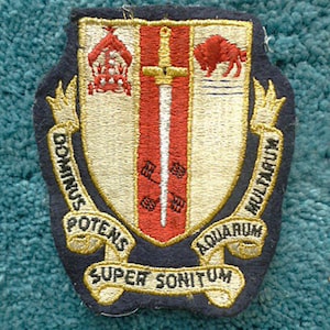 May include: A vintage embroidered patch with a shield design. The shield is divided into three sections. The left section features a red church with a white cross on top. The center section features a red sword with a white hilt. The right section features a red buffalo. The shield is surrounded by a gold scroll with the text "Potens Contrivus Aquarium Super Sonitum Multiplum".