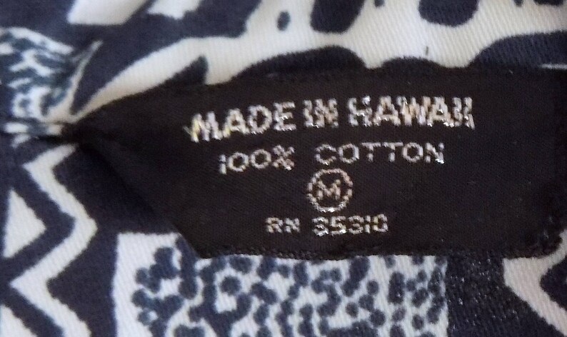 Pu&ograve; includere: Etichetta bianca e nera con il testo "Made in Hawaii 100% Cotton RN 35310" e una taglia "M" in un cerchio.