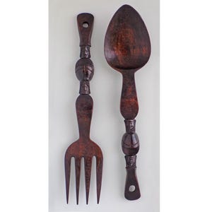 May include: A dark brown, carved wooden fork and spoon set. The handles feature carved faces and rounded shapes. The fork has four tines, and the spoon has a large, oval bowl. The set is designed for wall display.