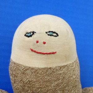 May include: A beige sock puppet with a brown knit bottom. The puppet has a hand-stitched face with blue eyes, a red nose, and a red smiling mouth.