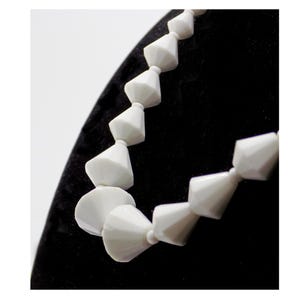 May include: A white necklace with geometric cone-shaped beads. The beads are strung together, creating a unique and modern design. The necklace is displayed against a black background.