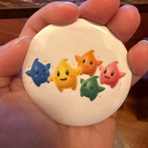 May include: A white button featuring five cartoon stars in blue, yellow, orange, green, and pink. Each star has a smiling face. The button is round and appears to be made of metal or plastic.