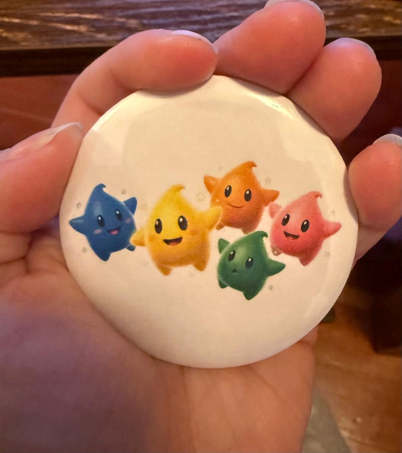 May include: A white button featuring five cartoon stars in blue, yellow, orange, green, and pink. Each star has a smiling face. The button is round and appears to be made of metal or plastic.