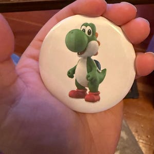 May include: A white button featuring a cartoon image of Yoshi, a green dinosaur character with a white belly, red shoes, and a blue shell. The button is round and has a glossy finish. The image is a vibrant and colorful illustration.