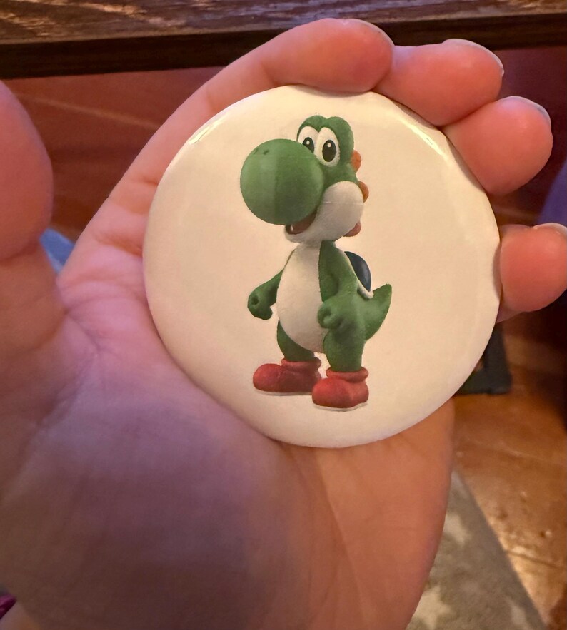 May include: A white button featuring a cartoon image of Yoshi, a green dinosaur character with a white belly, red shoes, and a blue shell. The button is round and has a glossy finish. The image is a vibrant and colorful illustration.