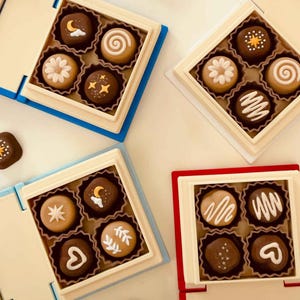 Puede incluir: Varias cajas de bombones con diversos diseños, incluyendo lunas, estrellas y espirales. Los bombones están dispuestos en pequeñas cajas cuadradas con interiores color crema y marcos coloridos azules y rojos. Algunos bombones están fuera de las cajas.