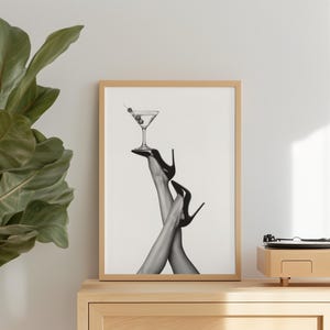Könnte beinhalten: Schwarz-Weiß-Druck mit einem Martini-Glas mit Oliven, das auf schwarzen High Heels balanciert. Der Druck wird auf einem Holzschrank angezeigt. Eine Pflanze und ein Plattenspieler sind ebenfalls sichtbar.