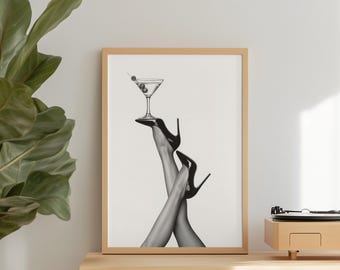 May include: Black and white framed print featuring a martini glass with olives balanced on high heels. The heels are black, and the print is displayed on a wooden cabinet. A plant and a record player are also visible.