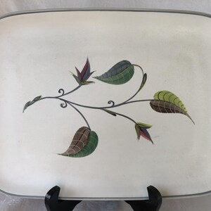 May include: A rectangular off-white serving platter with rounded corners and a thin grey rim. The centre features a stylised botanical illustration of a dark grey vine with green and brown patterned leaves and small purple and blue buds.