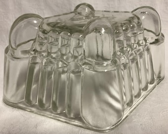 May include: A clear glass mold featuring a square base and a structured, multi-faceted design. The exterior surface is composed of vertical, rounded columns and four prominent, bulbous corner pillars that extend upward from the base.