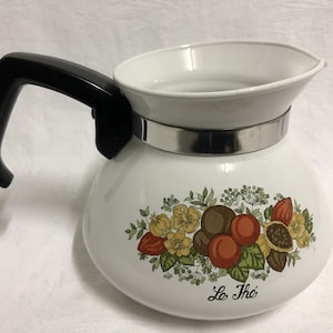 May include: A white enamel teapot with a black plastic handle and a silver-tone metal band around the neck. The body features a colourful botanical illustration of fruits and flowers with the text Le Thé printed in black script near the base.