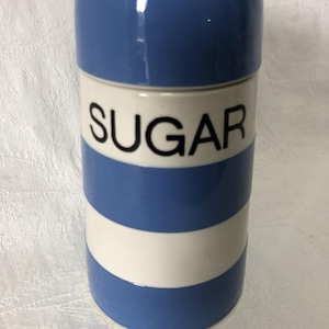 May include: A ceramic sugar container with a lid. The container is white with blue stripes and the word "SUGAR" in black. The lid is blue. The container is cylindrical in shape.