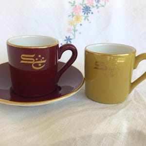 May include: A burgundy espresso cup with a matching saucer and a mustard yellow espresso cup. Both cups feature a gold-coloured logo with the letters SC and small star accents. The rims of the cups and the saucer are trimmed with a thin gold line.